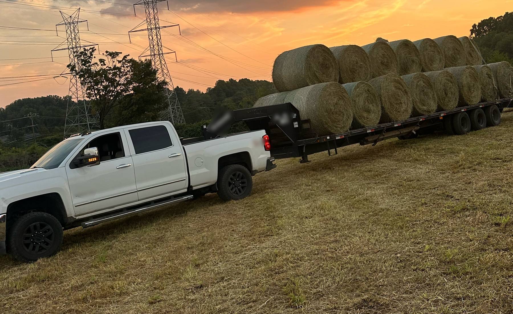 White pickup with a gooseneck trailer loaded with round hay bales in a mowed hayfield at sunset