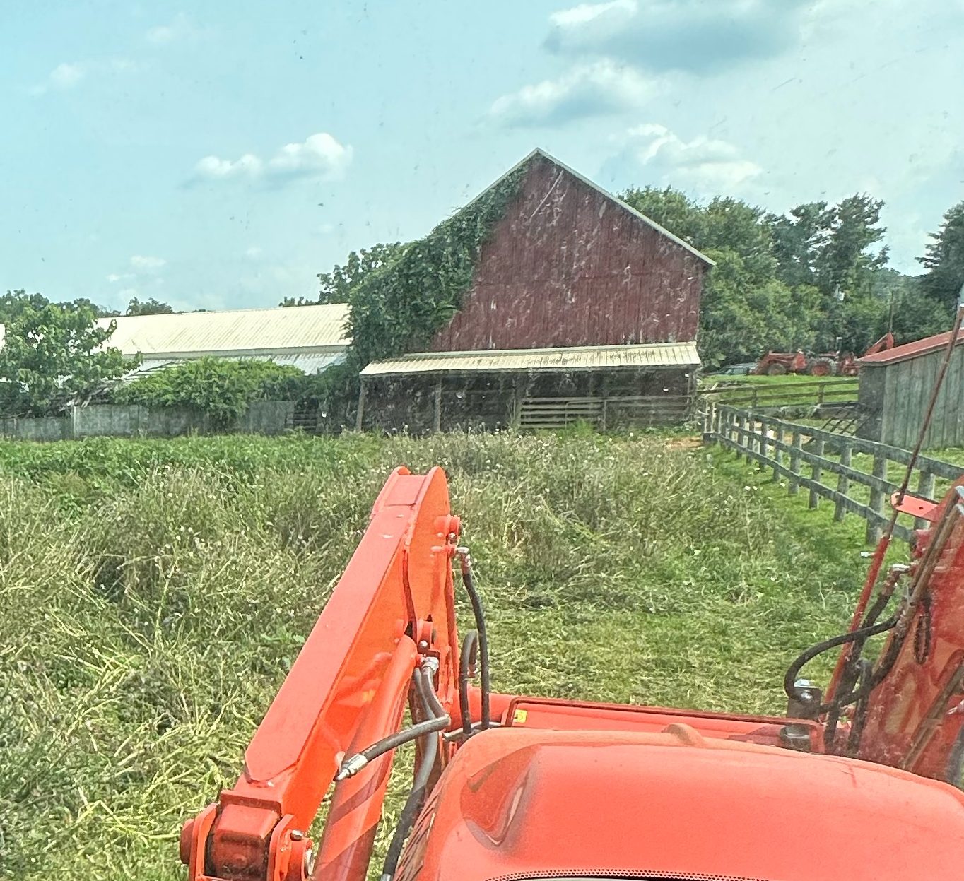 Tractor loader in an overgrown farm field near a red barn and board fence